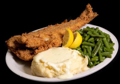 A plate featuring two fried fish fillets, mashed potatoes with gravy, green beans, and lemon wedges.