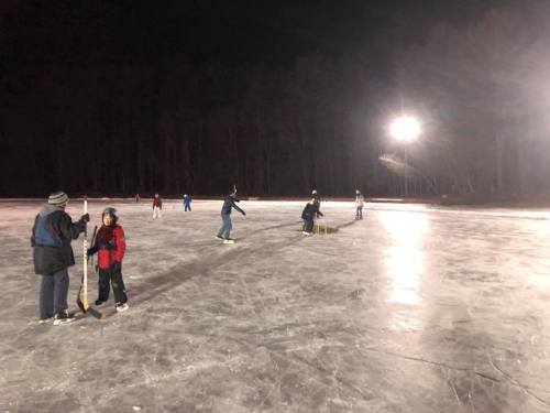 People ice skating on a frozen rink at night, with trees and lights in the background.