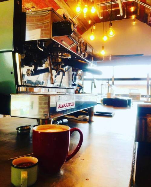 A coffee shop scene featuring a La Marzocco espresso machine and a red mug on a wooden table, with warm lighting.