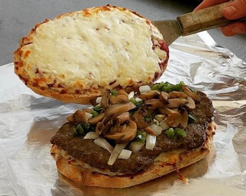 A hand lifts a cheesy pizza crust above a sandwich with a beef patty, mushrooms, and green peppers on a foil surface.