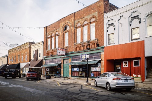 A quaint downtown street featuring brick buildings, shops, and parked cars under string lights.
