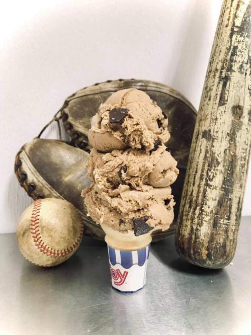 A tall ice cream cone with chocolate chunks, placed beside a baseball glove, ball, and bat on a metallic surface.