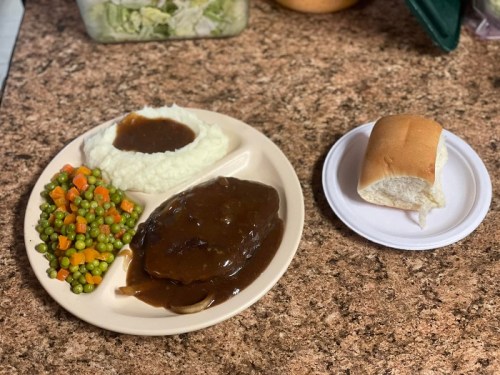 A plate with mashed potatoes, brown gravy, mixed vegetables, and sliced meat, alongside a dinner roll on a separate plate.