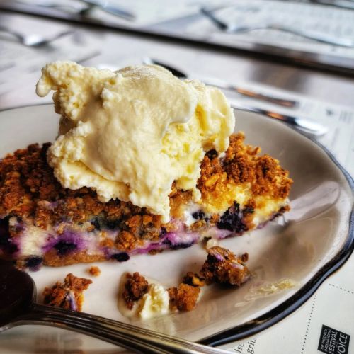 A slice of blueberry crumble topped with a scoop of vanilla ice cream on a plate, with a fork beside it.
