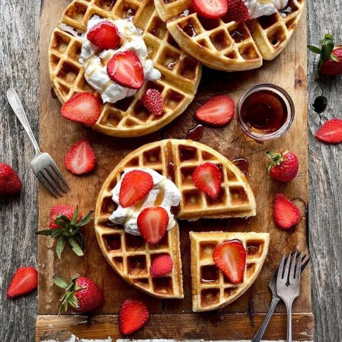 Three plates of golden waffles topped with strawberries and whipped cream, surrounded by fresh strawberries and syrup.