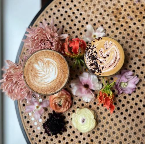 Two coffee drinks with decorative foam art, surrounded by colorful flowers on a textured wooden table.