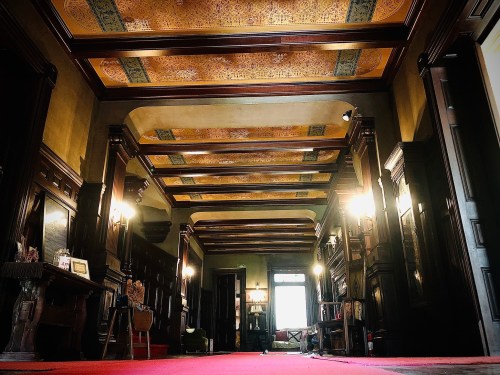 A grand hallway with ornate wooden beams, golden ceiling, and red carpet, illuminated by vintage wall sconces.