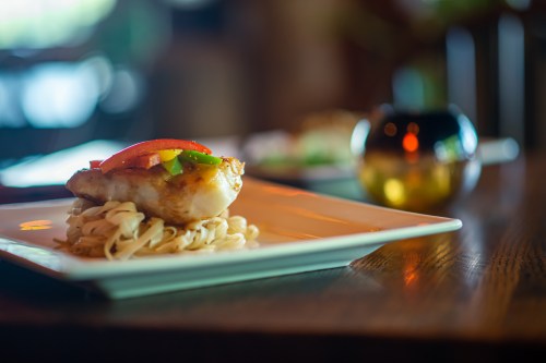 A plate of fish topped with peppers, served on a bed of noodles, with a softly lit background.