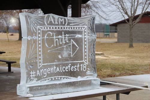 Ice sculpture with the text "Aim Chit" and hashtag "#ArgentIceFest18" displayed outdoors in a park setting.