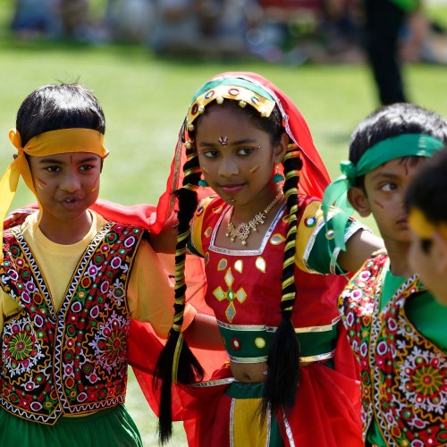 Children in colorful traditional outfits participate in a cultural performance outdoors, smiling and engaged.