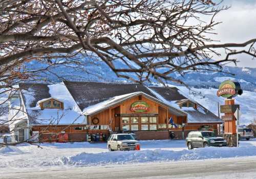 A cozy market building surrounded by snow, with mountains in the background and cars parked in front.