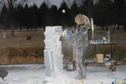 A person in a snowy landscape carves ice, wearing protective gear and a furry hat, with tools and equipment nearby.