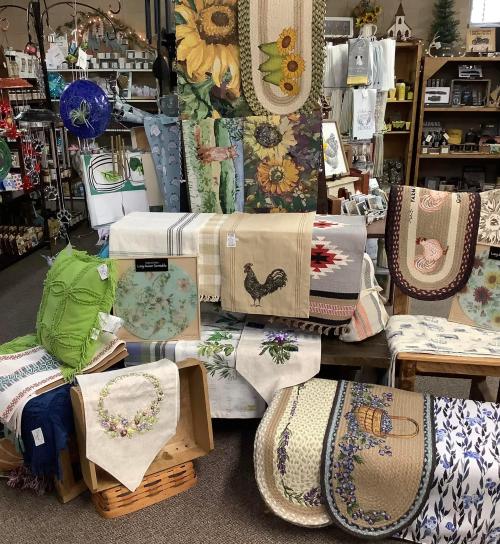 A display of various decorative table linens and home textiles featuring floral and animal designs in a shop setting.