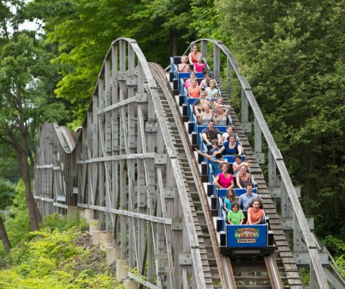 A wooden roller coaster with excited riders going up a steep incline surrounded by lush greenery.