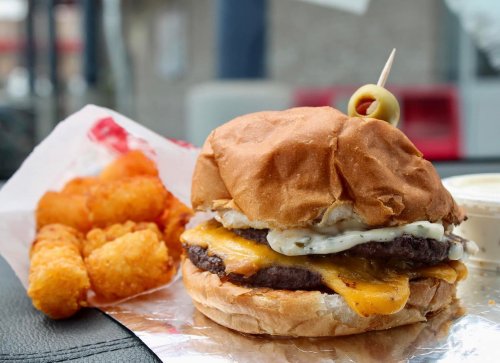 A close-up of a cheeseburger with a pickle on top, served with crispy tater tots on the side.