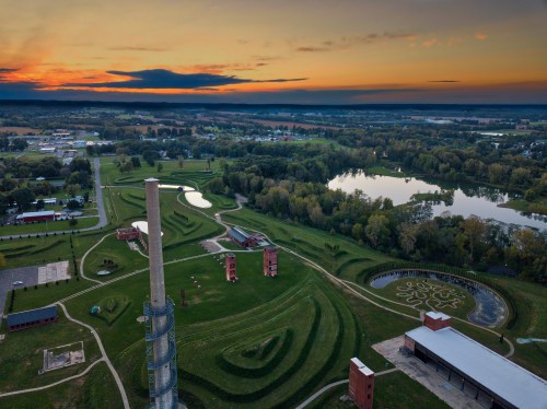 Aerial view of a park with winding paths, a tall tower, and a serene lake at sunset.