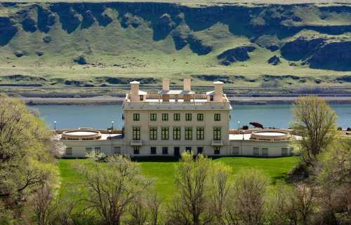 A large, elegant building with a manicured lawn, set against a backdrop of green hills and a river.