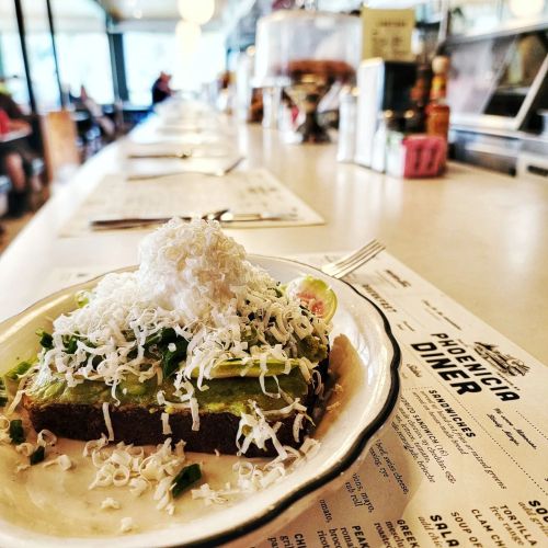 A plate of avocado toast topped with shredded cheese, served at a diner with a long counter in the background.