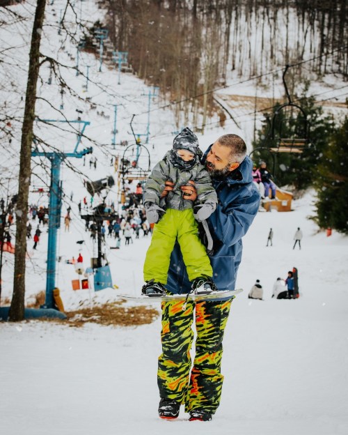 A man holds a child in winter gear on a snowy slope, with skiers and a ski lift in the background.