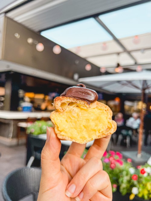 A hand holds a pastry filled with cream and topped with chocolate, set against a blurred outdoor café background.