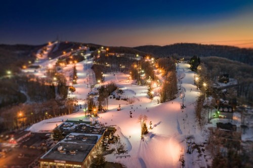 A snowy ski resort at dusk, with illuminated slopes and a cozy lodge surrounded by trees.
