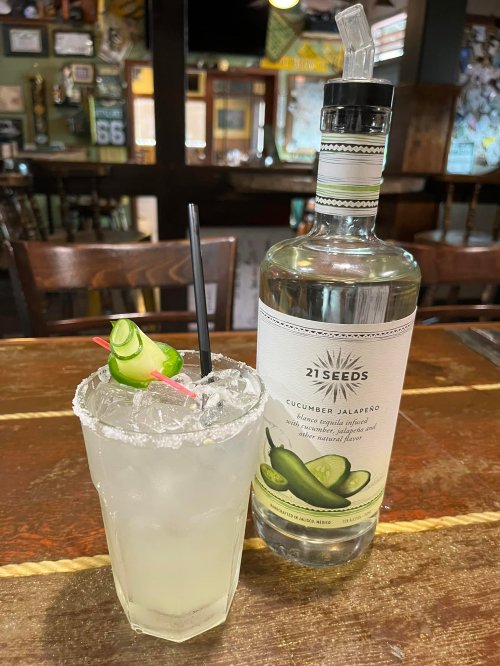 A refreshing cucumber jalapeño cocktail with a salted rim, garnished with cucumber, next to a bottle of 21 Seeds.