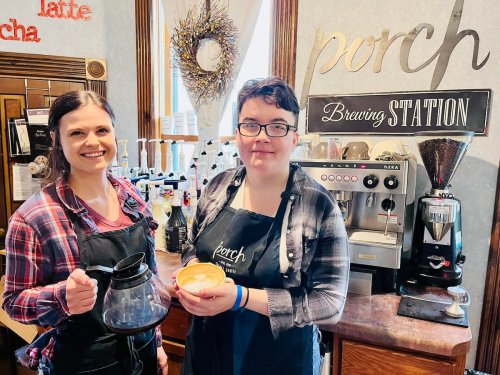 Two baristas stand in a coffee shop, one holding a coffee pot and the other a latte, with brewing equipment in the background.