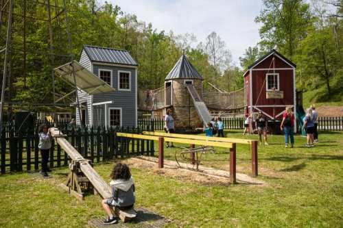 Children and adults play in a park with colorful buildings, a playground, and green trees in the background.
