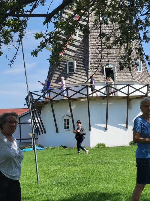 A group of people walking on a balcony of a windmill, with green grass and trees in the foreground.