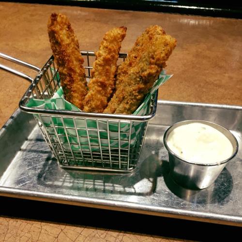 A small metal basket with crispy fried sticks and a cup of dipping sauce on a metal tray.