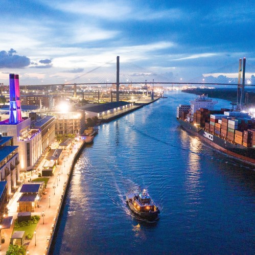 Aerial view of a river with a boat, city lights, and a bridge under a twilight sky.