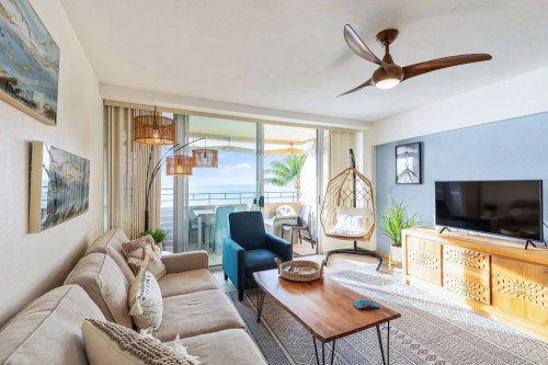 Bright, modern living room with a sofa, coffee table, TV, and a hanging chair, featuring large windows and ocean views.