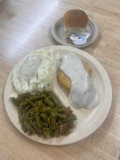 A plate with fried chicken smothered in gravy, mashed potatoes, green beans, and a roll with butter on the side.