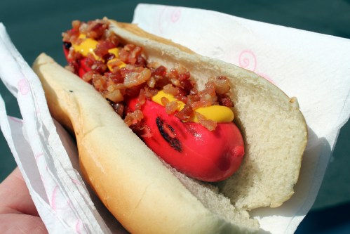 A hot dog in a bun topped with mustard and crispy bacon bits, held in a hand with a light background.