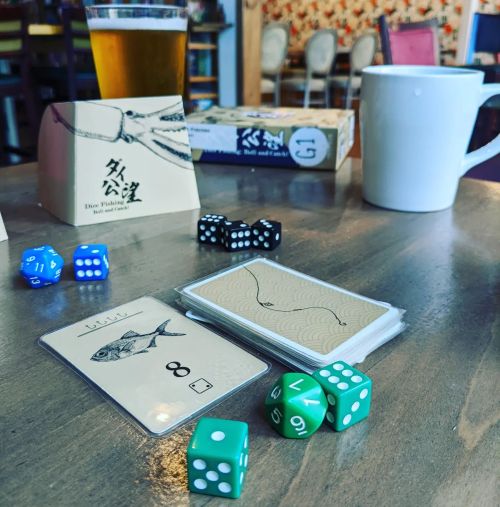 A tabletop scene featuring a beer, a coffee mug, dice, and cards from a board game in a cozy setting.