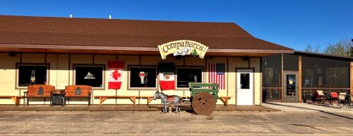 A rustic building with flags, benches, and a sign reading "Companeros" under a clear blue sky.