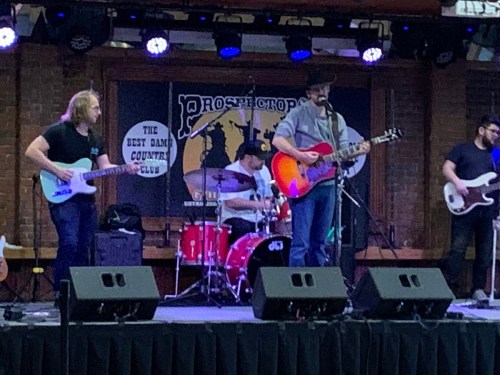 A band performs on stage with guitars and drums, set against a backdrop featuring a country club logo.