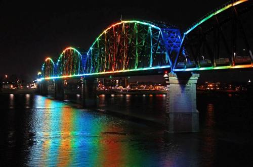 A brightly lit bridge arches over a river at night, reflecting vibrant rainbow colors on the water's surface.
