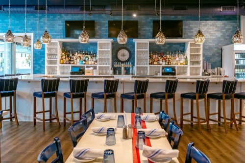 A modern restaurant interior featuring a bar with high stools, a dining area with tables, and blue tiled walls.