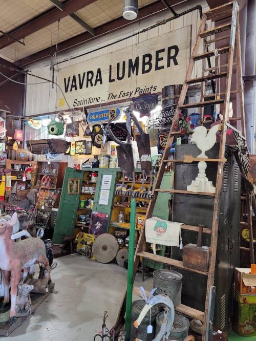 A vintage shop display featuring a "Vavra Lumber" sign, ladders, and various antique items and signs.