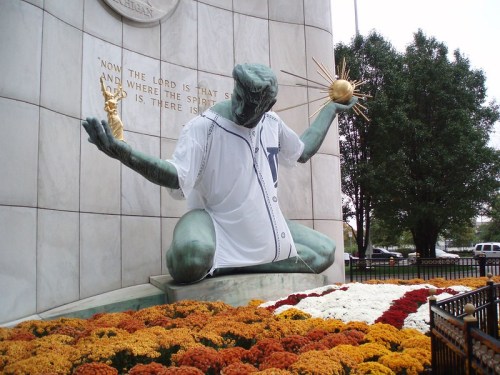 A statue of a figure in a baseball jersey, surrounded by colorful flowers, in front of a stone monument.