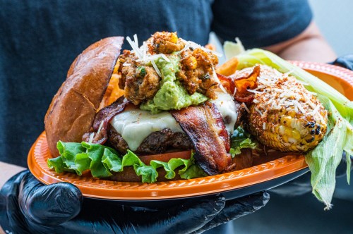 A gourmet burger topped with bacon, guacamole, and crispy onions, served with grilled corn on the side.