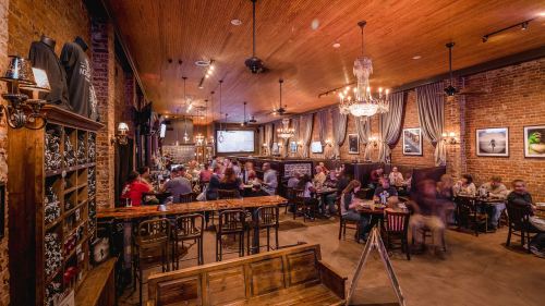 A lively restaurant interior with wooden decor, chandeliers, and patrons enjoying meals at tables.