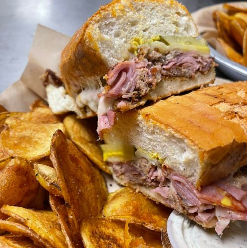 A close-up of a sandwich filled with meats and pickles, served with crispy potato chips on the side.
