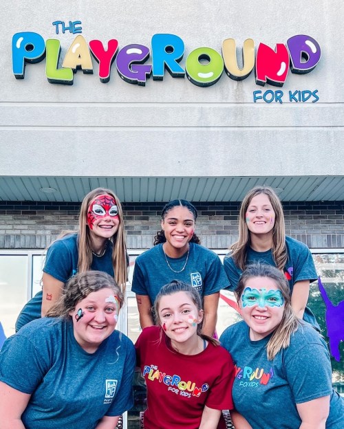 A group of six smiling women with face paint, posing in front of a colorful sign that reads "The Playground for Kids."