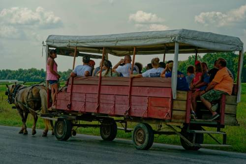 A horse-drawn wagon filled with people rides along a rural road under a cloudy sky.