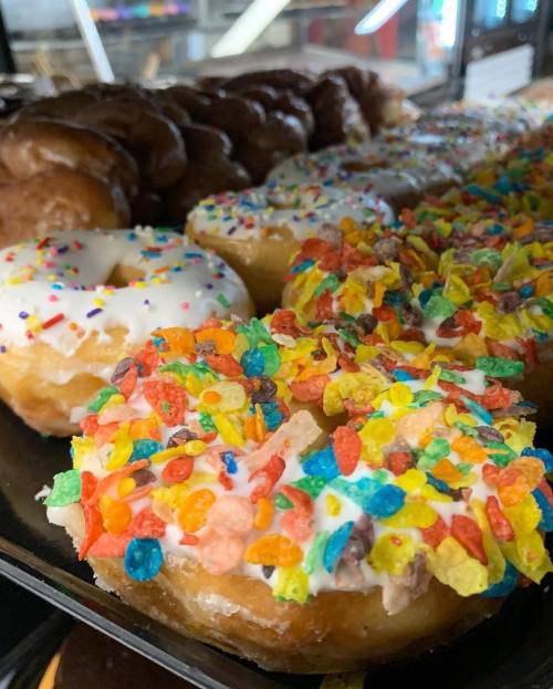 A display of colorful donuts, including one topped with frosting and fruity cereal, alongside glazed and chocolate varieties.