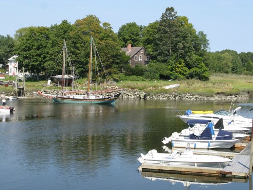 A calm harbor scene with a tall ship and several small boats docked along the shore, surrounded by greenery.