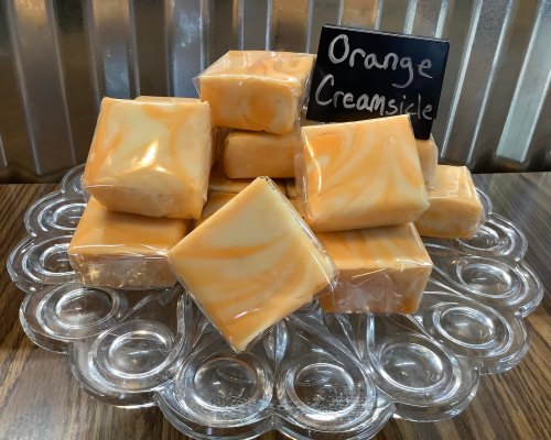 A clear dish holds stacked orange creamsicle soap bars, wrapped in plastic, with a small sign labeling the scent.
