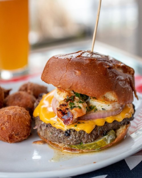 A close-up of a cheeseburger with shrimp, served with golden-brown fritters and a glass of beer in the background.